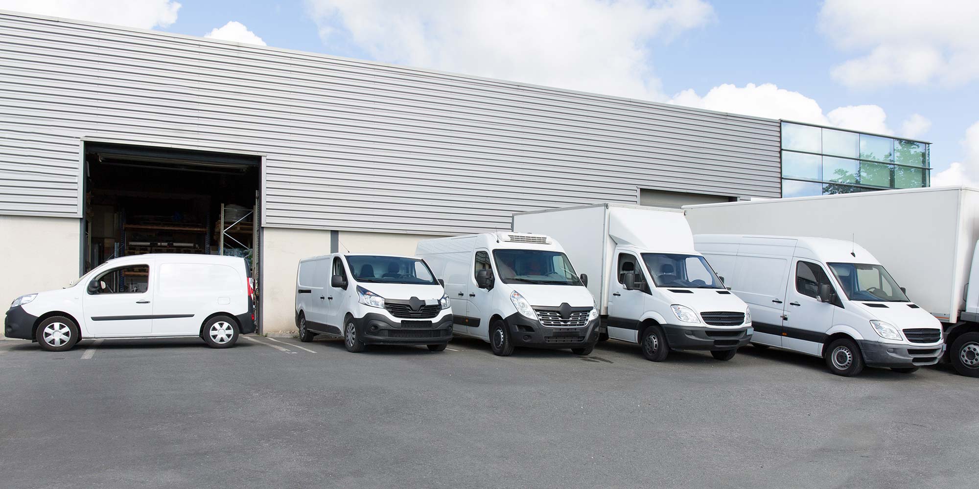Five white delivery vans parked outside a large warehouse with a metal exterior.