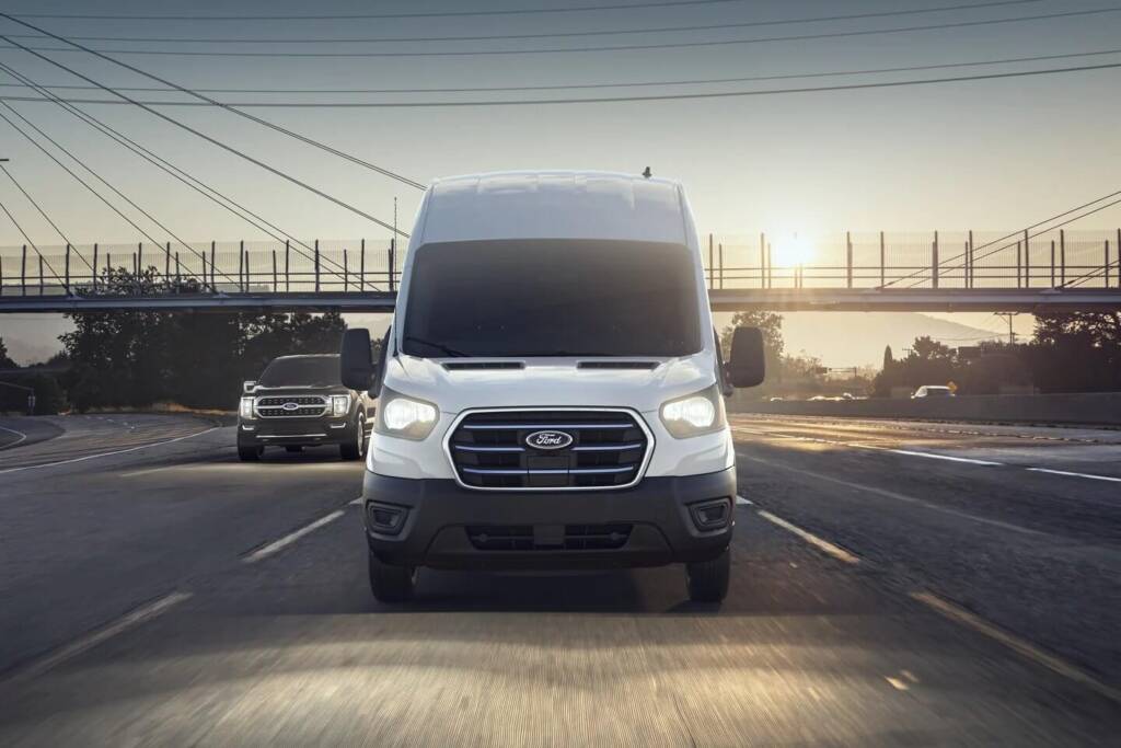 White van driving on highway at sunrise with a bridge and another vehicle in the background.