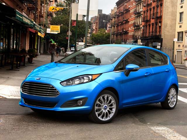A blue Ford Fiesta parked on a city street with brick buildings and storefronts in the background.