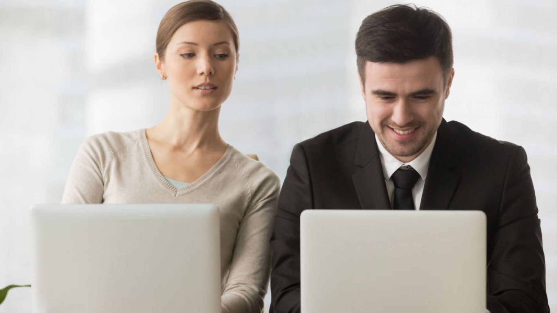 Two professionals working on laptops at a desk, one in a suit smiling, the other in a sweater with a neutral expression.