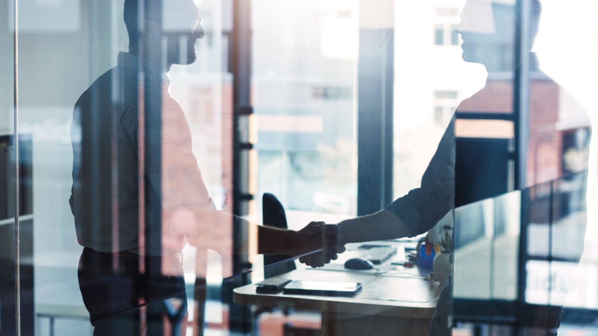 Two people shaking hands in an office, visible through glass reflections.