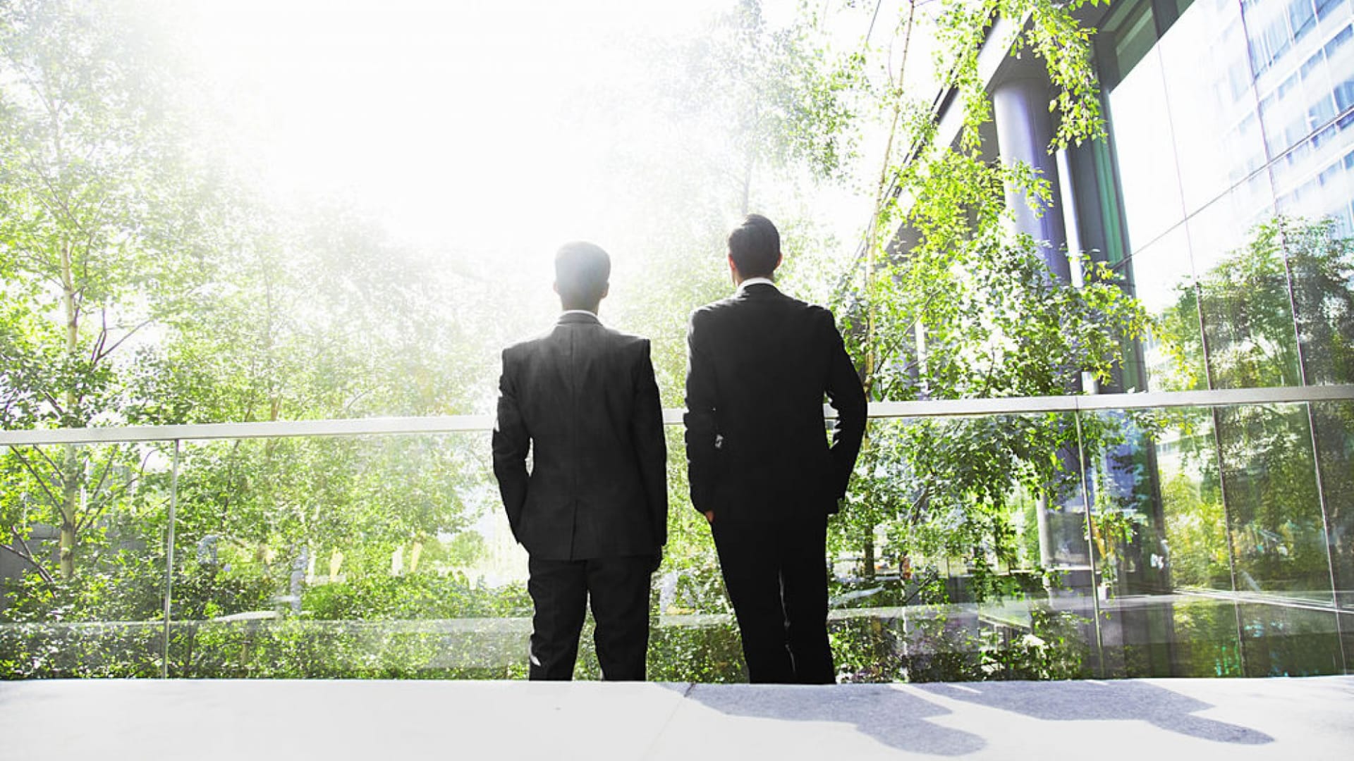 Two people in suits stand on a balcony overlooking a green, sunlit garden with glass buildings in the background.