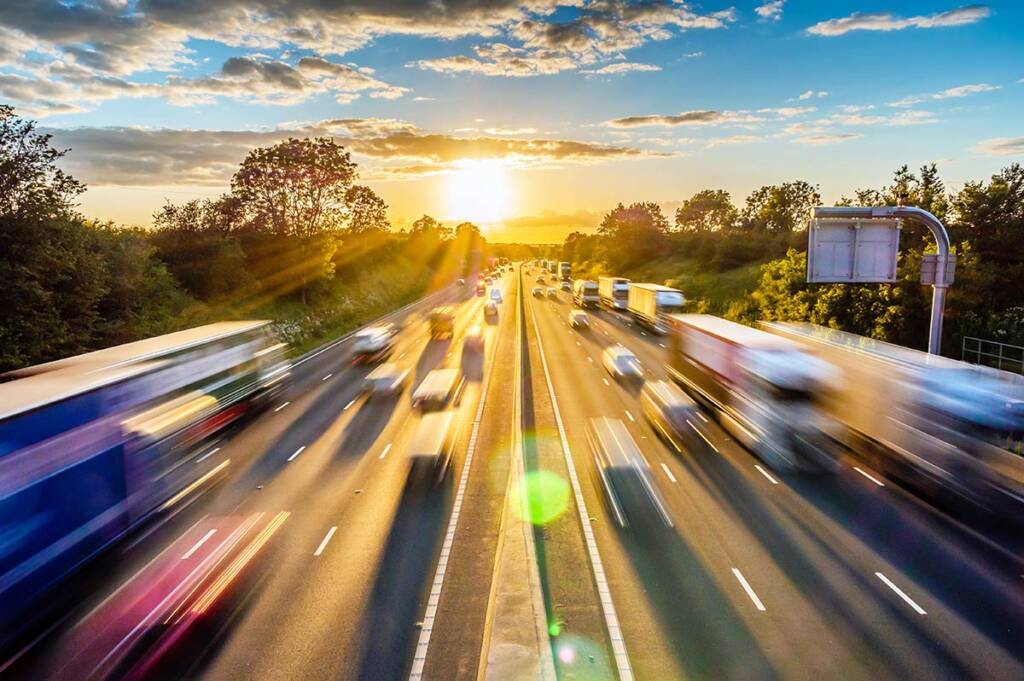 Blurred highway traffic at sunset, with trees on both sides and a vibrant sky with clouds.