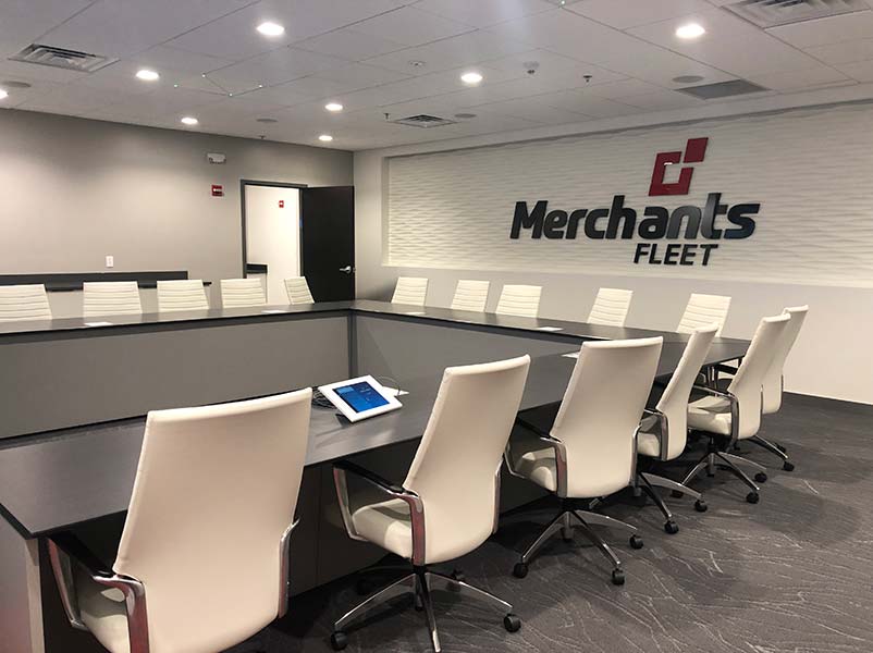 Conference room with a long table, white chairs, and a tablet on the table. "Merchants Fleet" logo on the wall.