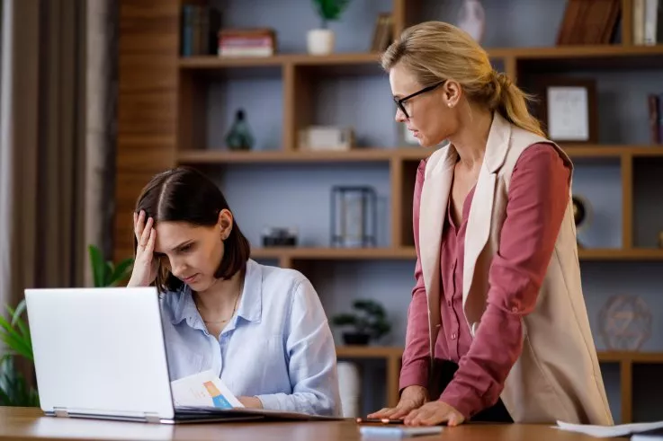 Woman in blue shirt looks frustrated at a laptop while another woman in glasses leans over to assist her.