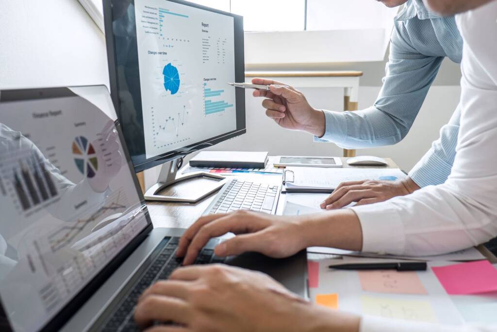 Two people work at a desk with laptops, analyzing graphs and charts on computer screens.