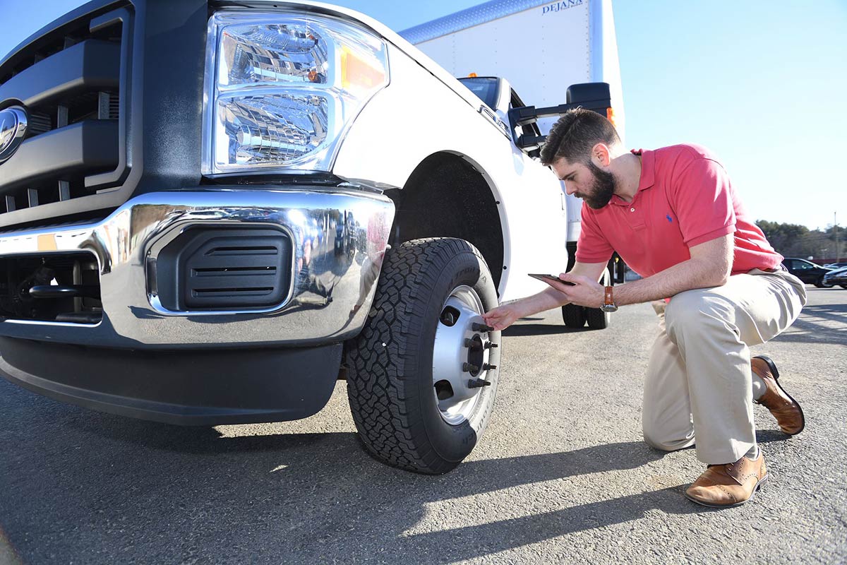 Man in red shirt inspecting a white truck's front wheel in a sunny parking lot.