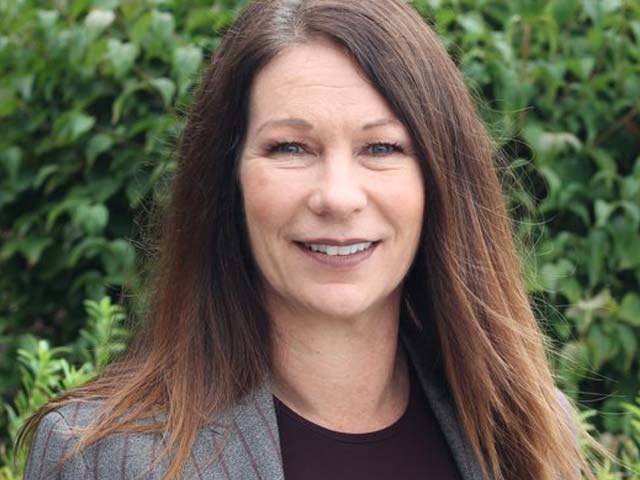 Smiling woman with long brown hair standing outside, wearing a blazer, with green foliage in the background.