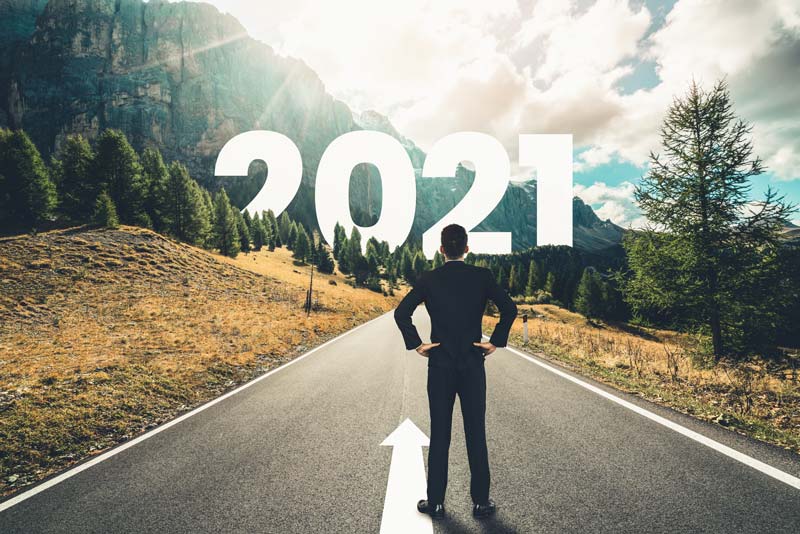 A person in a suit stands on a road facing large "2021" text amidst mountains and trees under a bright sky.