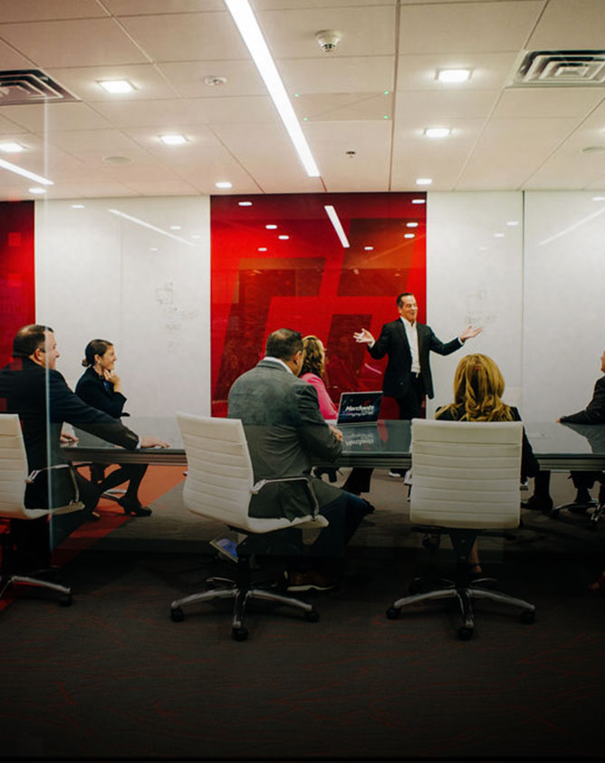 A person stands gesturing in front of a seated group in a modern conference room with red and white decor.