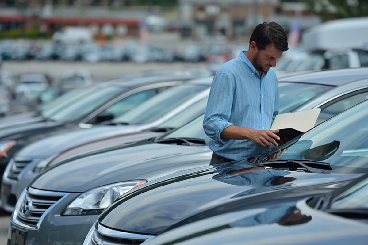 Man in blue shirt holding a folder, inspecting parked cars in a dealership lot.