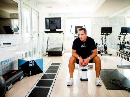 A man sitting on a bench in a well-lit gym with exercise equipment around him.