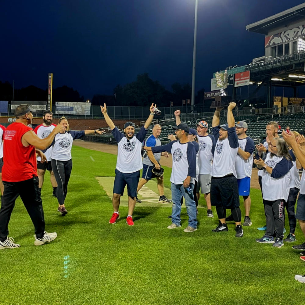 A team celebrates on a baseball field at night, with arms raised and smiles, as one person gives a thumbs up.