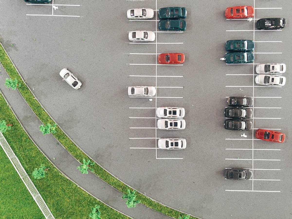 Aerial view of a parking lot with parked cars and a single car driving on the road beside the greenery.