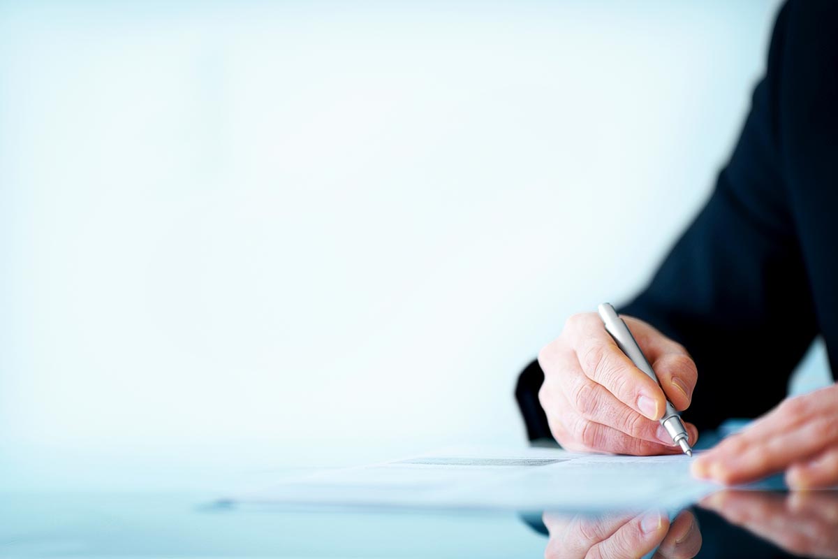 A person in a suit signs a document with a pen on a reflective table.