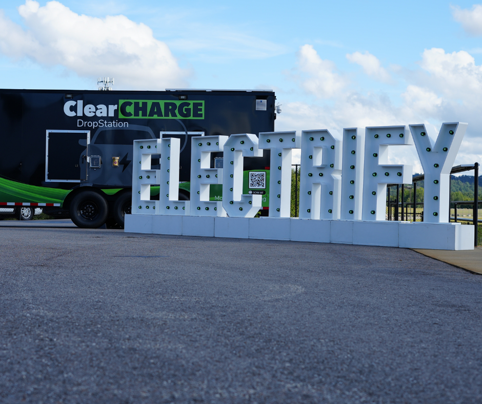 Large "ELECTRIFY" sign and ClearCharge DropStation trailer on a paved surface, with cloudy sky background.