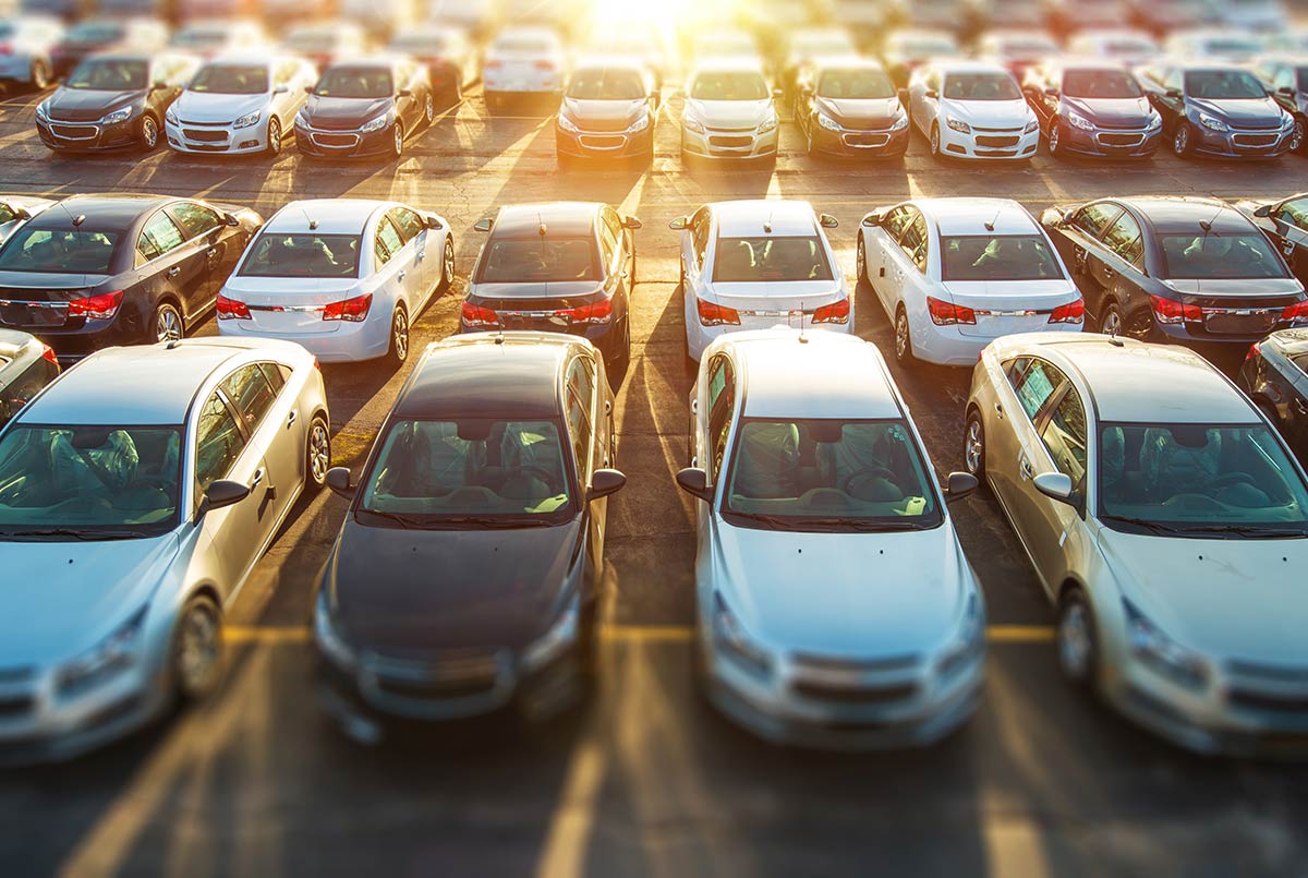 Aerial view of a parking lot filled with rows of cars under a bright sun.