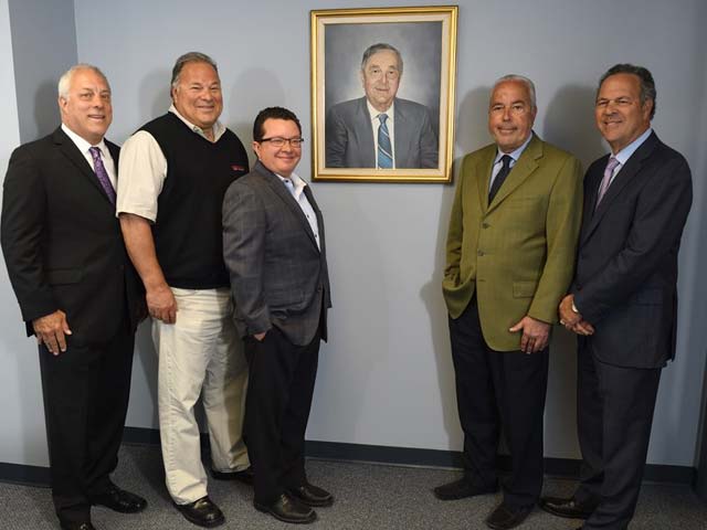 Five men standing together in an office, smiling at the camera, with a portrait on the wall behind them.