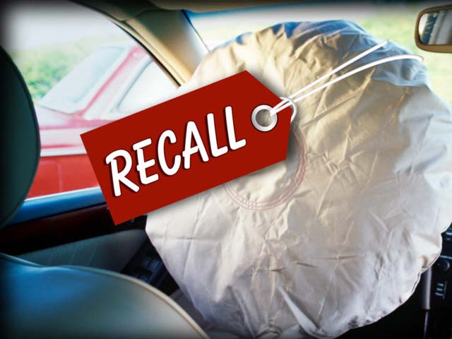 An inflated airbag in a car with a red "RECALL" tag attached.