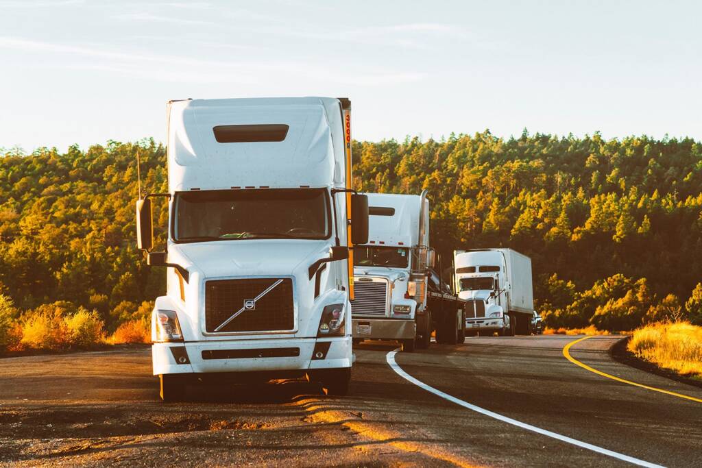Three white semi-trucks parked on a winding road against a backdrop of trees and clear sky.