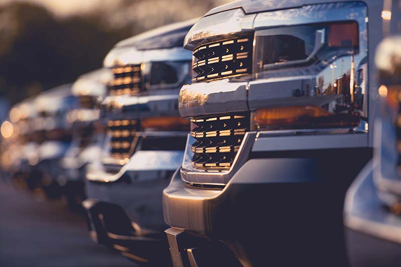 Close-up of a row of parked trucks with gleaming chrome grilles reflecting light at sunset.