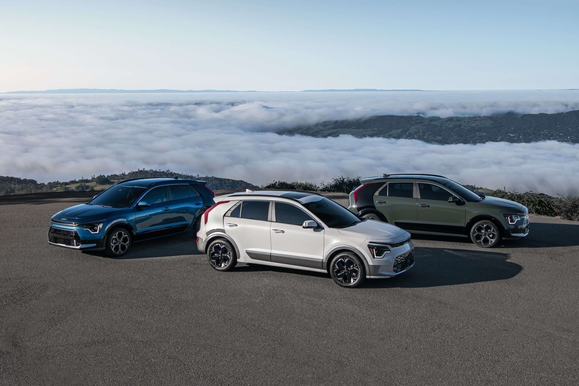 Three cars parked on a scenic overlook above clouds with hills in the background.