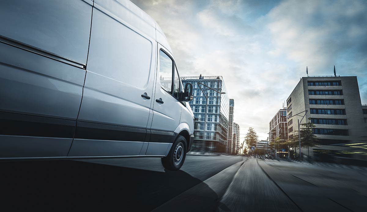 White van driving through a modern urban area with tall buildings and a dynamic, blurred motion effect.