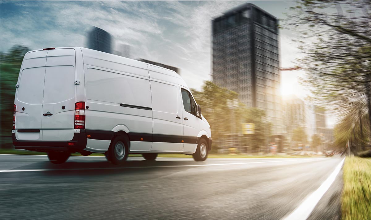 White delivery van driving fast on a city street with blurred buildings and trees in the background.