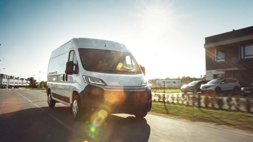 White delivery van driving on a sunny road with a building in the background.