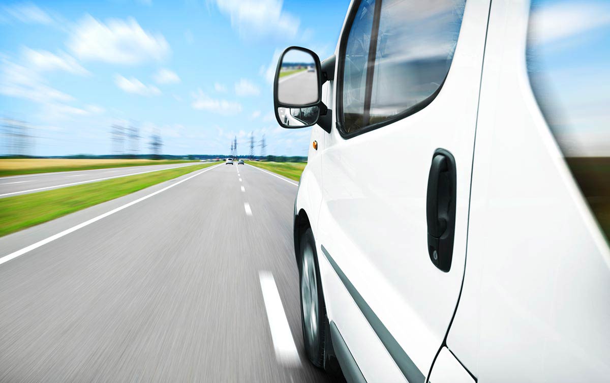 White van speeding down a highway under a clear blue sky with distant trees and vehicles.