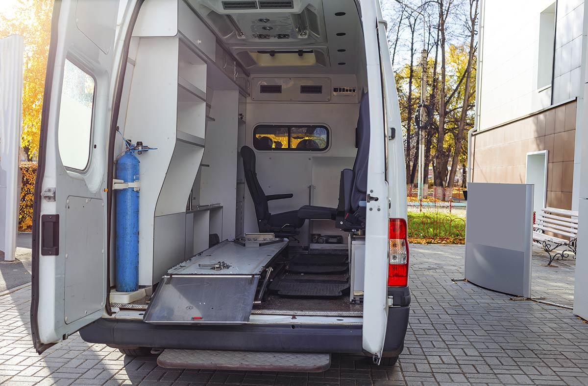 Interior of a converted van with a blue oxygen cylinder and folded stretcher, parked on a paved area.