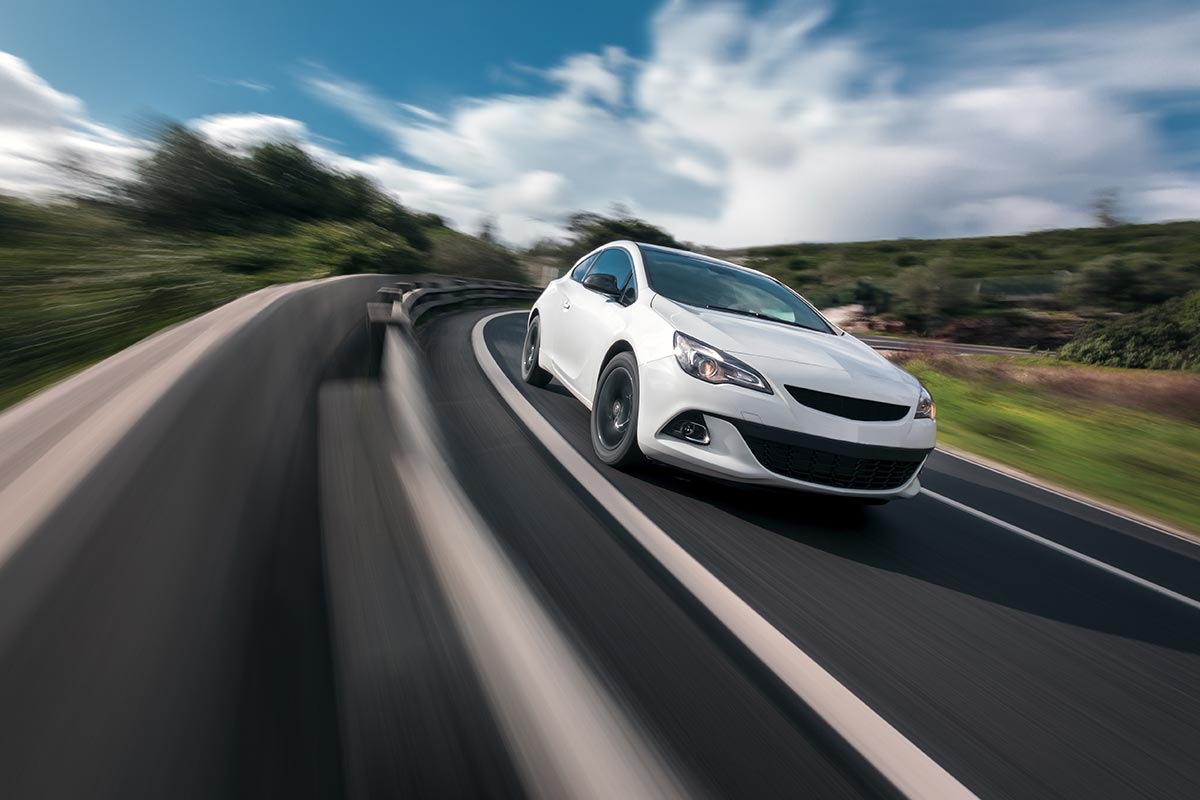 White car speeding on a curvy road, with motion blur in the background, blue sky, and greenery on the sides.