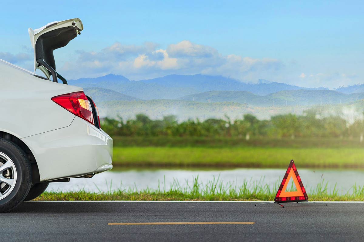 Car with open trunk parked on roadside, warning triangle placed behind, mountain landscape in background.
