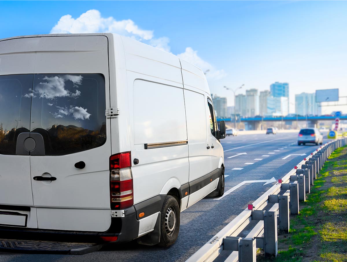 White delivery van parked on the side of a highway, city skyline in the background under a clear blue sky.