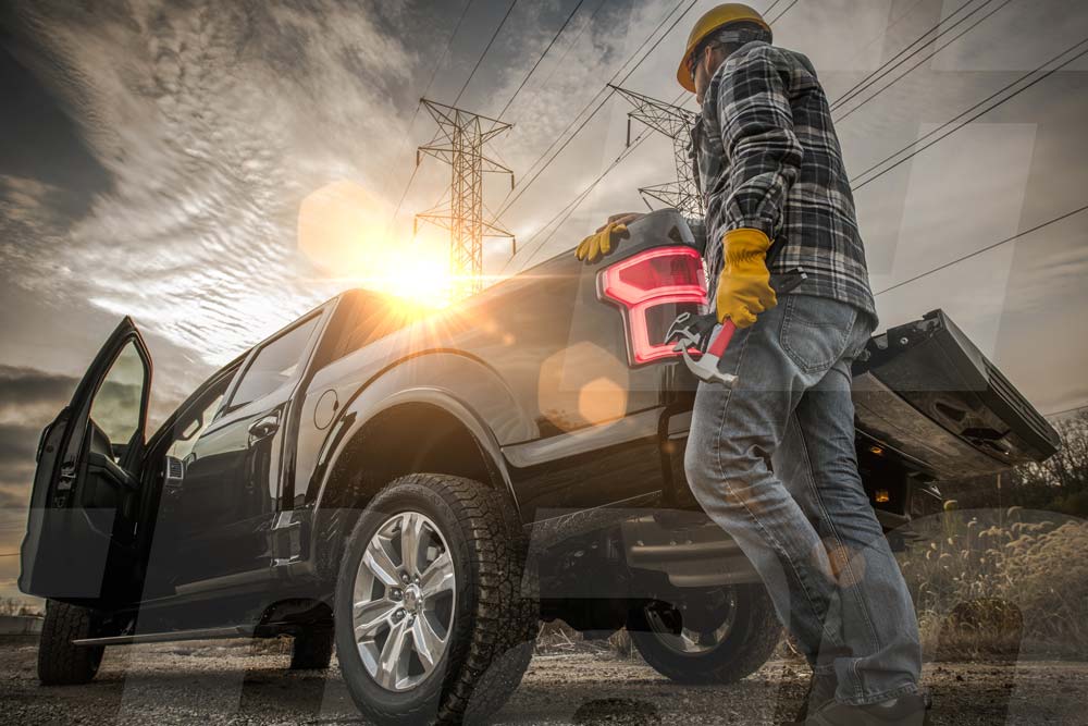 Worker in safety gear stands next to a pickup truck under power lines at sunset, holding tools.
