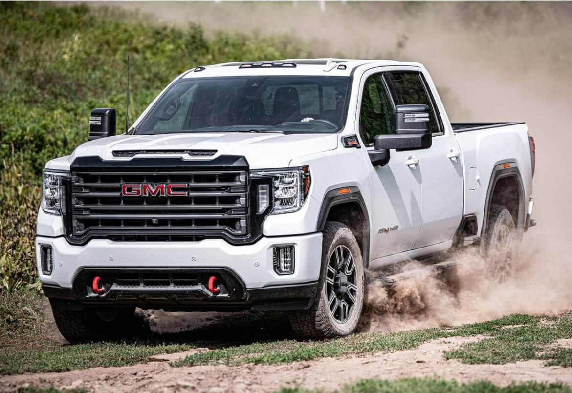 A white GMC pickup truck driving off-road on a dusty trail with green foliage in the background.