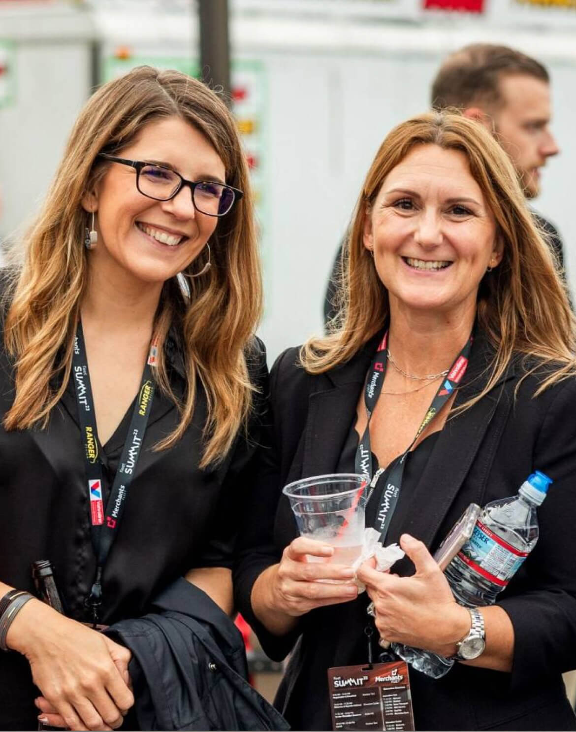 Two women smiling, wearing black outfits and event badges, holding drinks at an outdoor event.