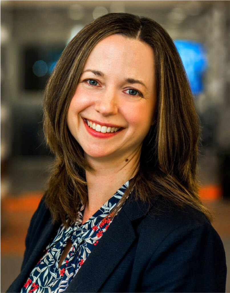 Smiling woman with long brown hair wearing a blue patterned blouse and dark blazer, blurred background.