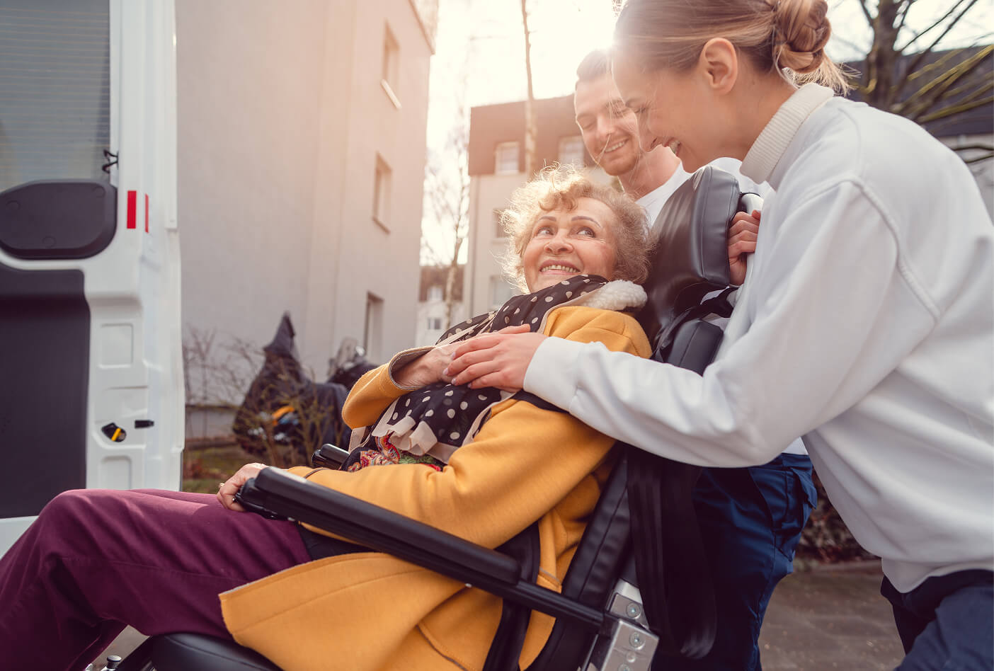Elderly person in a wheelchair smiles while two caregivers assist them outdoors.