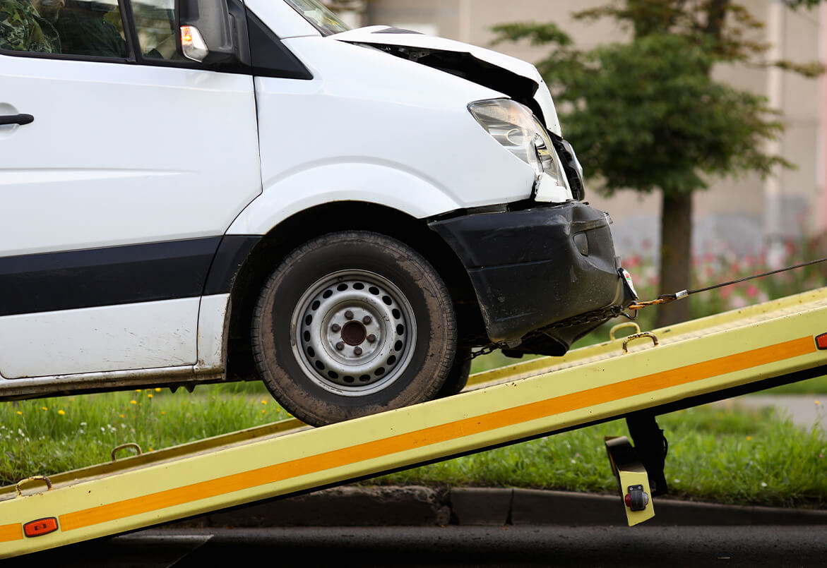 Van with front damage being loaded onto a tow truck ramp, with grass and trees in the background.