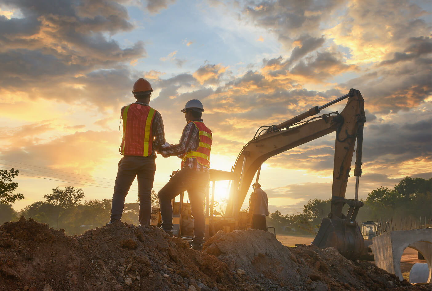 Two construction workers in safety gear stand near an excavator on a dirt mound at sunset.