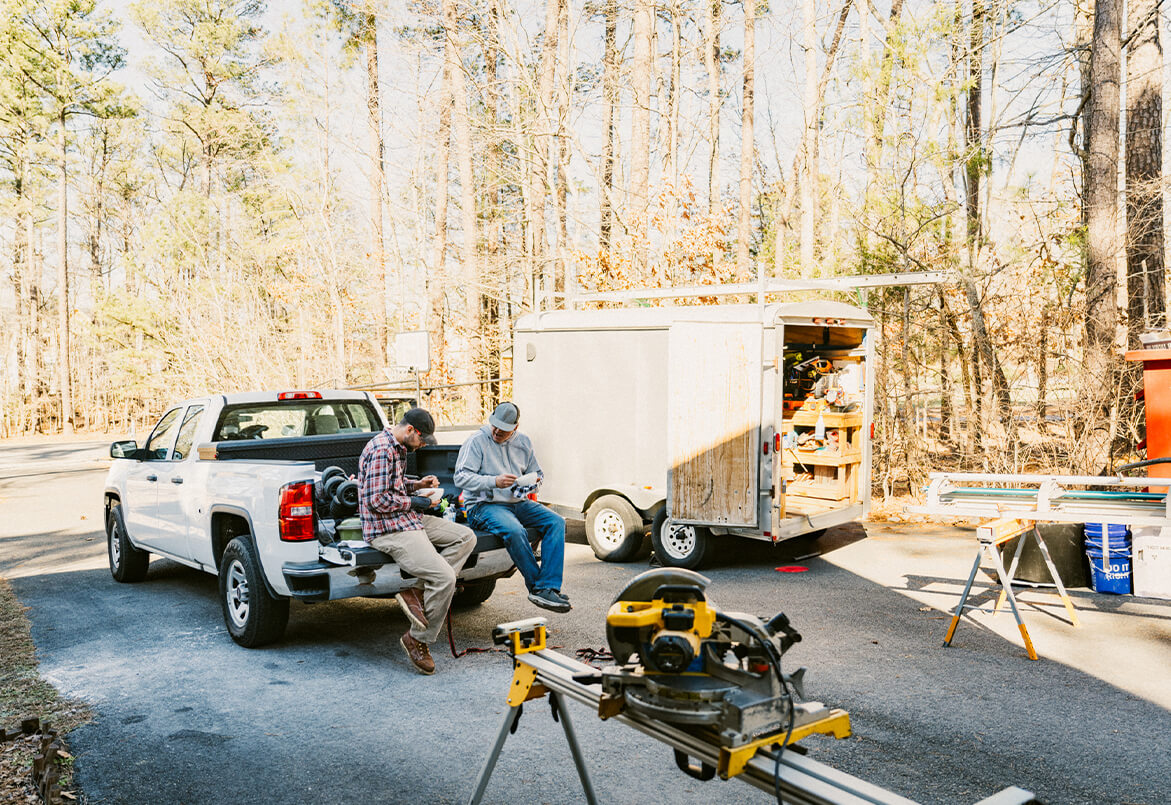 Two people sitting on a pickup truck's tailgate, near a trailer with tools, in a sunny wooded area.