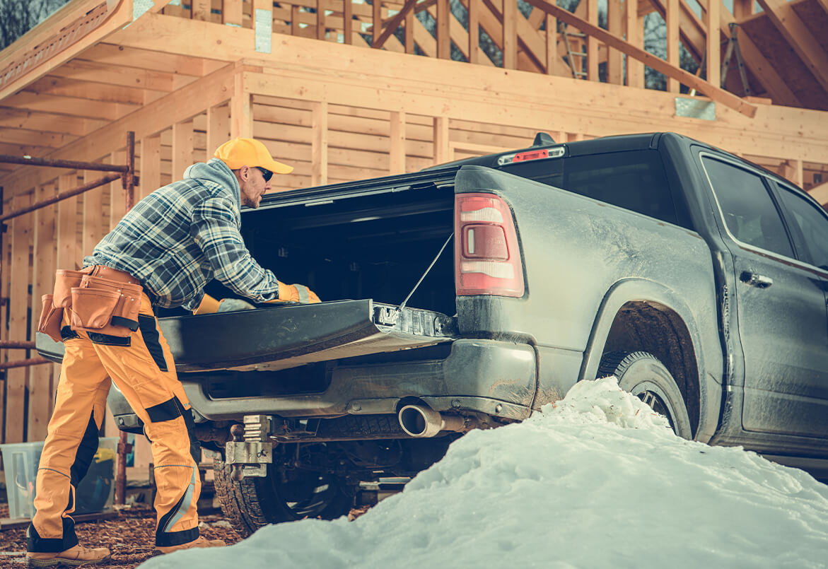 Worker in yellow and plaid, unloading tools from a pickup truck near a snowy construction site.