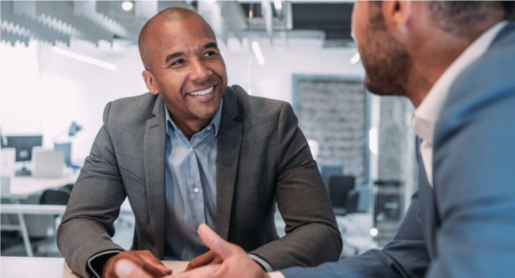 Two professionals in suits talking and smiling at a table in a modern office setting.