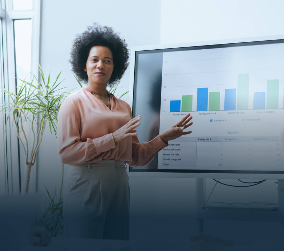 Person presenting data on a screen with bar graphs and tables, standing next to a potted plant.