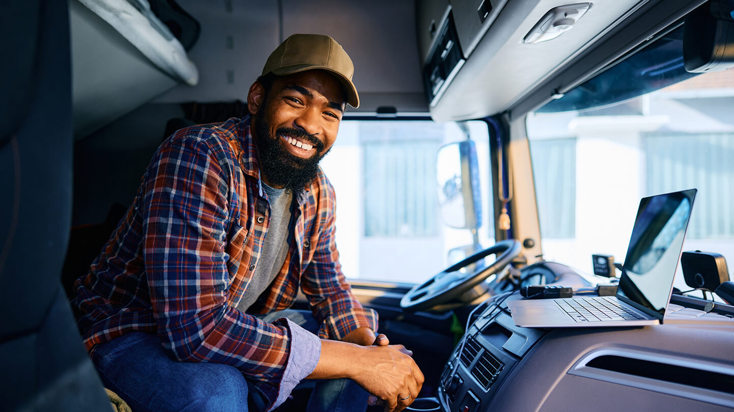 Smiling man in a plaid shirt and cap sitting inside a truck cabin with a laptop on the dashboard.