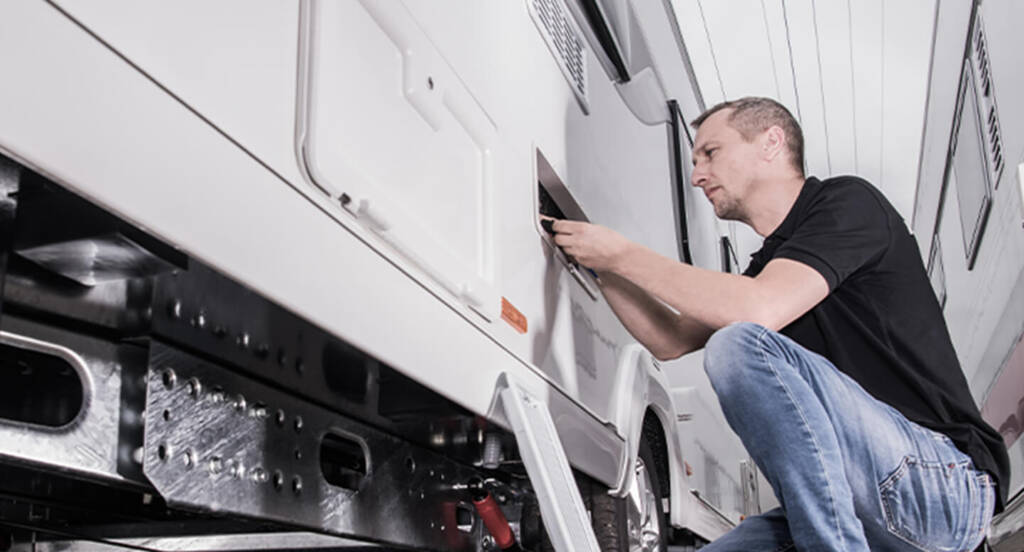 Man in a black shirt and jeans working on the side panel of a white RV in a garage.