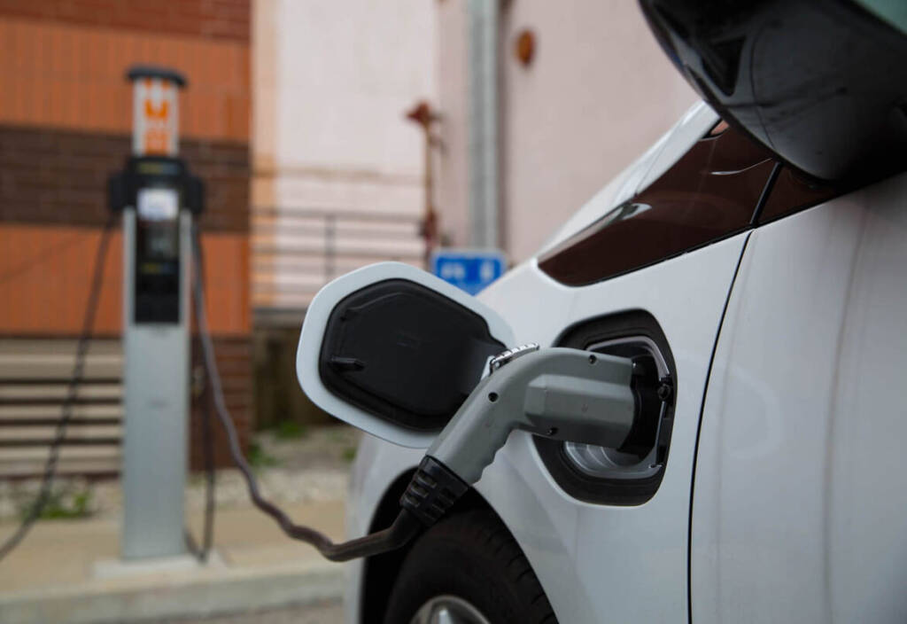 Close-up of a white electric vehicle charging at a public charging station with a brick wall in the background.