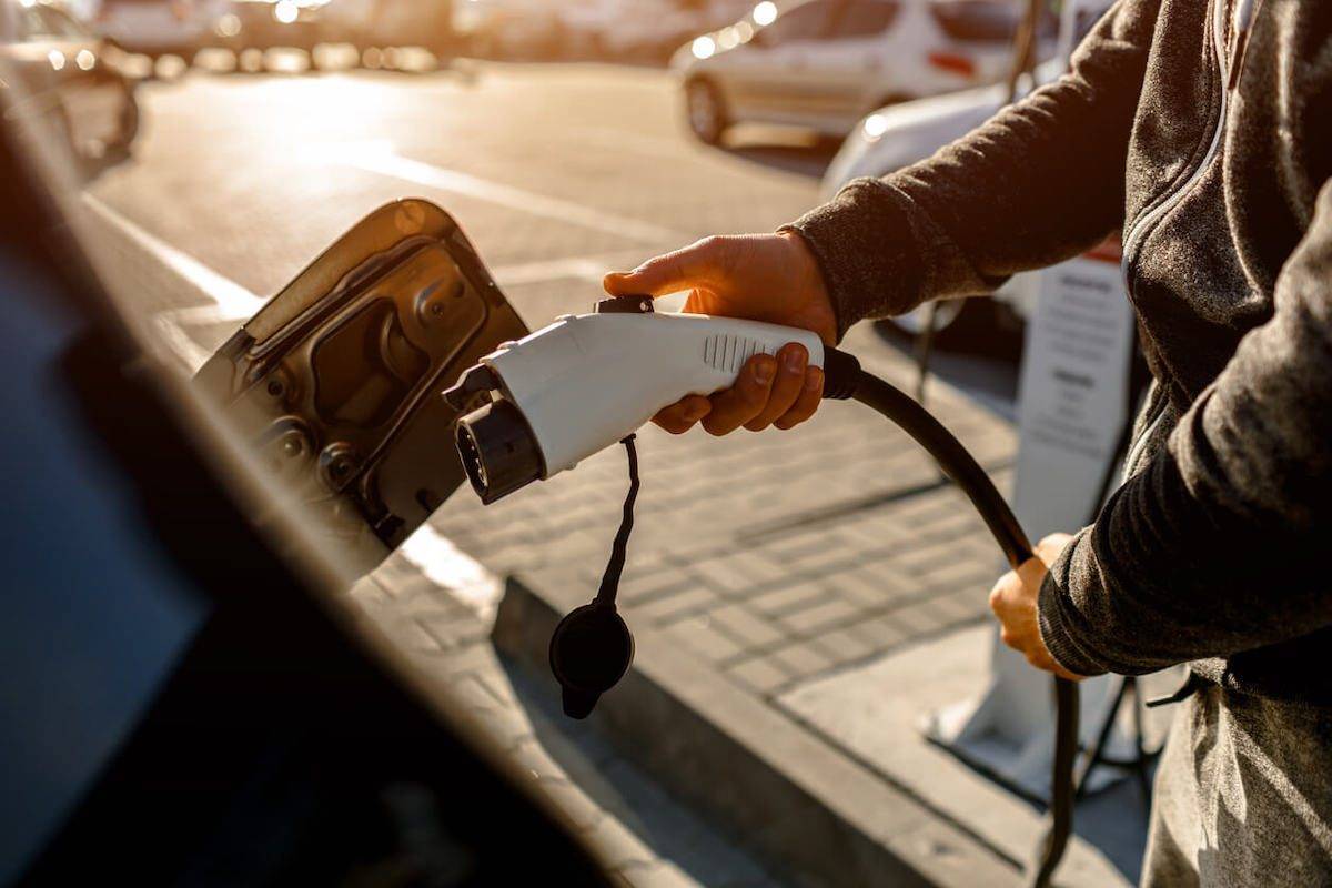 Person plugging an electric car charger into a vehicle at sunset in a parking lot.