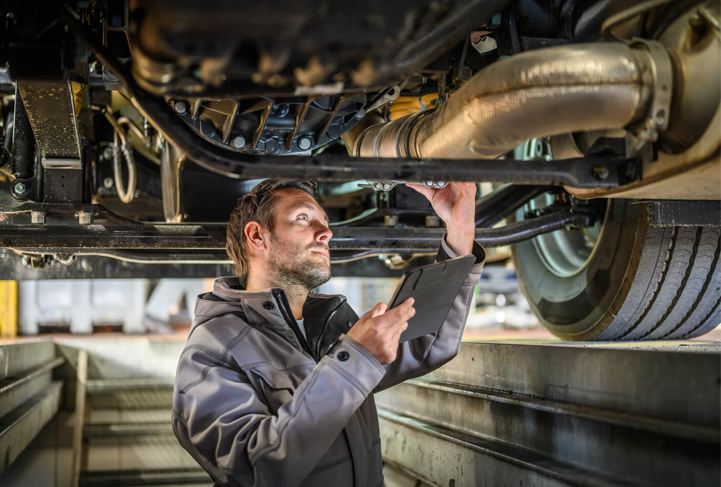Mechanic inspecting a vehicle's undercarriage while holding a tablet in a garage setting.
